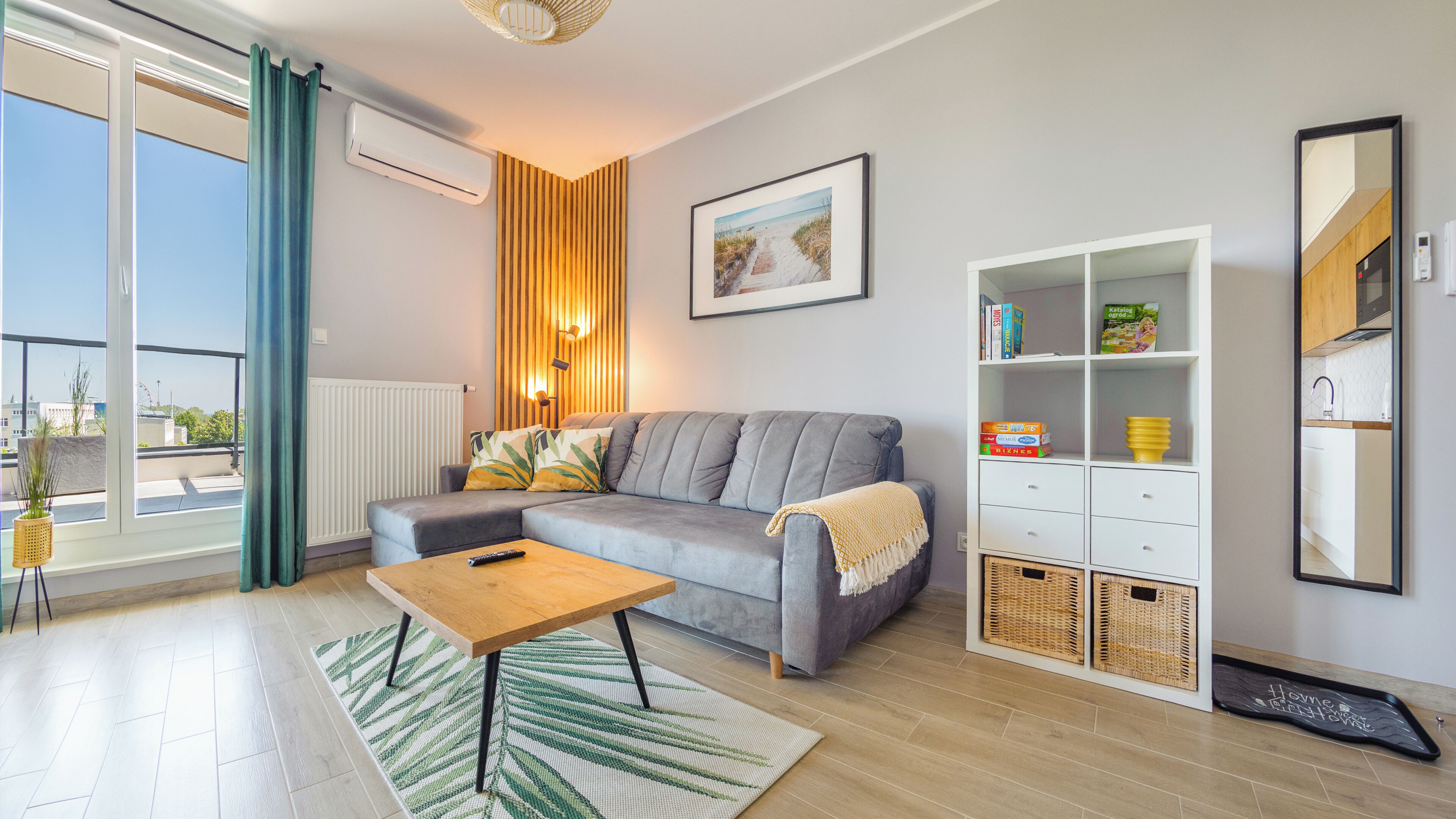 A living room with a grey sofa, a wooden coffee table, a green and white rug, a wall-mounted air conditioner, a framed picture, and a white cabinet with shelves.