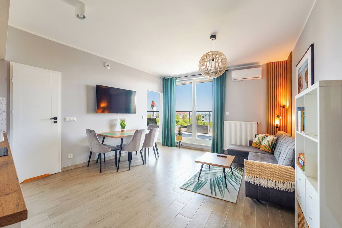 A modern living room with a sofa, a coffee table, a dining area with chairs, a TV mounted on the wall, and a balcony with curtains.