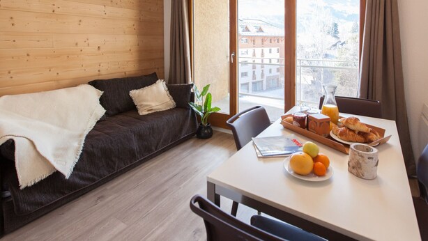 Une pièce chaleureuse avec un mur en bois, un canapé sombre agrémenté d’un plaid blanc, une table à manger où sont disposés des aliments pour le petit-déjeuner, et une vue sur des bâtiments et des montagnes, visible à travers de grandes fenêtres.
