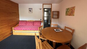 A compact room with a wooden floor, a small sofa with a pink patterned cover, a wooden dining table with chairs, and a small wooden cabinet.