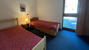 A room with two single beds, a small table with a lamp, a window with curtains, and a view of a parking area.