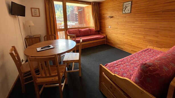 A room with a wooden wall, a bed, a sofa, a table with chairs, a TV, and a window with curtains.