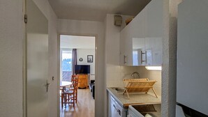 A narrow kitchen with a stove, sink, and cabinets. A wooden chair and table are in the adjacent dining area.
