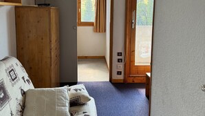 Une pièce avec une armoire en bois, un téléviseur fixé au mur, un canapé à motifs et, vue par une porte, un espace extérieur doté d’une porte et d’une fenêtre en bois.