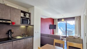 A compact kitchen with a microwave, kettle, and kettle on the countertop. There is a dining area with a table and chairs, and a sofa. A TV is mounted on the wall, and a window offers a view of a mountainous landscape. - Ski-in apartment with pool and parking (Flaine)