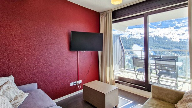 A modern living room with a red wall, a flat-screen TV mounted above a wooden coffee table, and a large window offering a mountain view. - Ski-in apartment with pool and parking (Flaine)