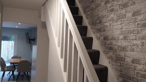 A staircase with white balusters and dark carpeting, leading to a room with a brick wall and a dining area in the background. - 7 Eyre Square Lane (Galway)