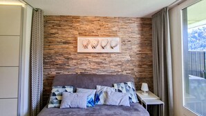 A bedroom with a stone wall, a bed with patterned pillows, a bedside table with a lamp, and a window with a view. - Renovated studio in Flaine center with balcony for 5 people (Flaine)