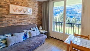 A modern living room with a stone wall, a grey sofa with patterned cushions, a small table with a lamp, and a balcony with a scenic view. - Renovated studio in Flaine center with balcony for 5 people (Flaine)