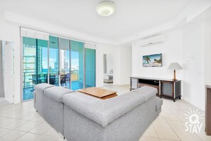 A modern living room with a grey sofa, a wooden coffee table, a TV stand, and a sliding glass door leading to a balcony with a city view. - Family Getaway at Wings Resort 3 Bedroom Apartment (Surfers Paradise)