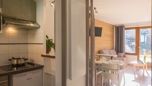 Une cuisine moderne dotée d’une cuisinière intégrée et d’un lave-vaisselle compact. Un coin repas avec une table et des chaises est visible à travers une porte ouverte, donnant sur un salon avec un canapé et une fenêtre.