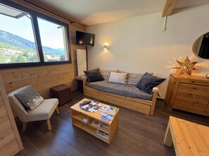 A cozy room with a wooden sofa, a small table with books, a chair, and a TV mounted on the wall.