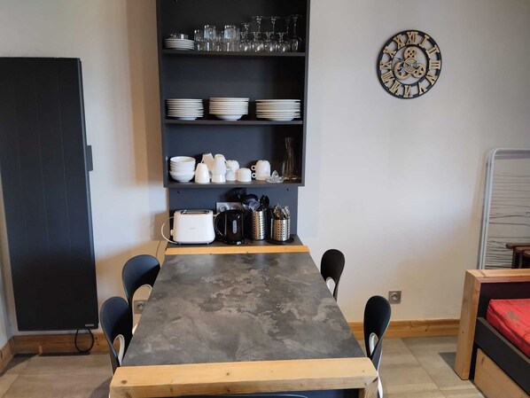 A dining area with a table, chairs, a black cabinet with dishes and glasses, a clock, and a bed with a red mattress. - Comfortable apartment with free parking in La Plagne (La Plagne Montchavin Les Coches)