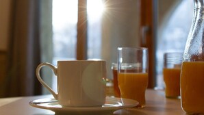 Una escena de desayuno con una taza y platillo blancos, un vaso de jugo de naranja y una jarra de jugo de naranja sobre una mesa junto a una ventana.