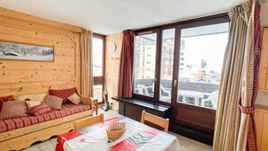Une pièce chaleureuse avec un canapé en bois, une table à manger agrémentée d’un panier, et une grande fenêtre avec des rideaux.