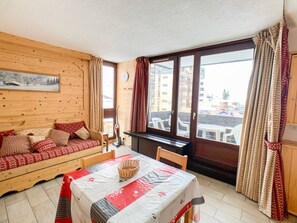 A cozy room with a wooden sofa, a dining table with a basket, and a large window with curtains.
