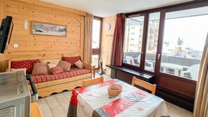 Une pièce chaleureuse avec un mur en bois, un canapé orné de coussins rouges, une table recouverte d’une nappe blanche et une fenêtre agrémentée de rideaux.