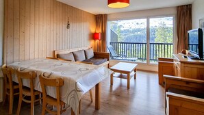 Quarto com piso de madeira, uma cama com cabeceira, uma mesa com cadeiras, uma mesinha auxiliar, uma TV sobre um suporte de madeira e uma varanda com vista para árvores e colinas.