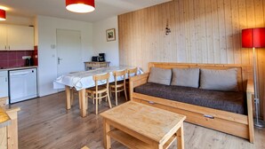 Um espaço de estar compacto com parede de madeira, um sofá, uma mesa de jantar com cadeiras, uma kitchenette e um abajur vermelho.