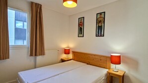 Quarto com cama de casal, duas luminárias de cabeceira, janela com cortinas e dois quadros na parede.