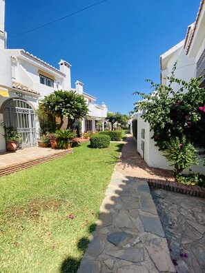 A narrow stone pathway flanked by white buildings with tiled roofs, leading to a grassy area with potted plants and a small bush.