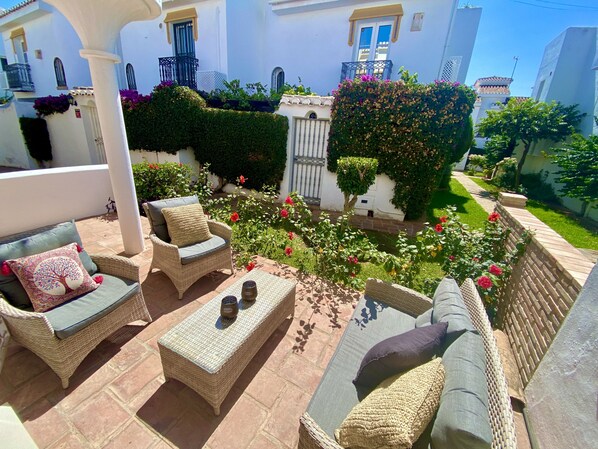 A sunlit outdoor patio with wicker furniture, a small table, and a garden with flowering plants.