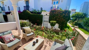 Eine sonnige Terrassengrundstück mit Korbmöbeln, einem kleinen Tisch und einem Garten mit blühenden Pflanzen.