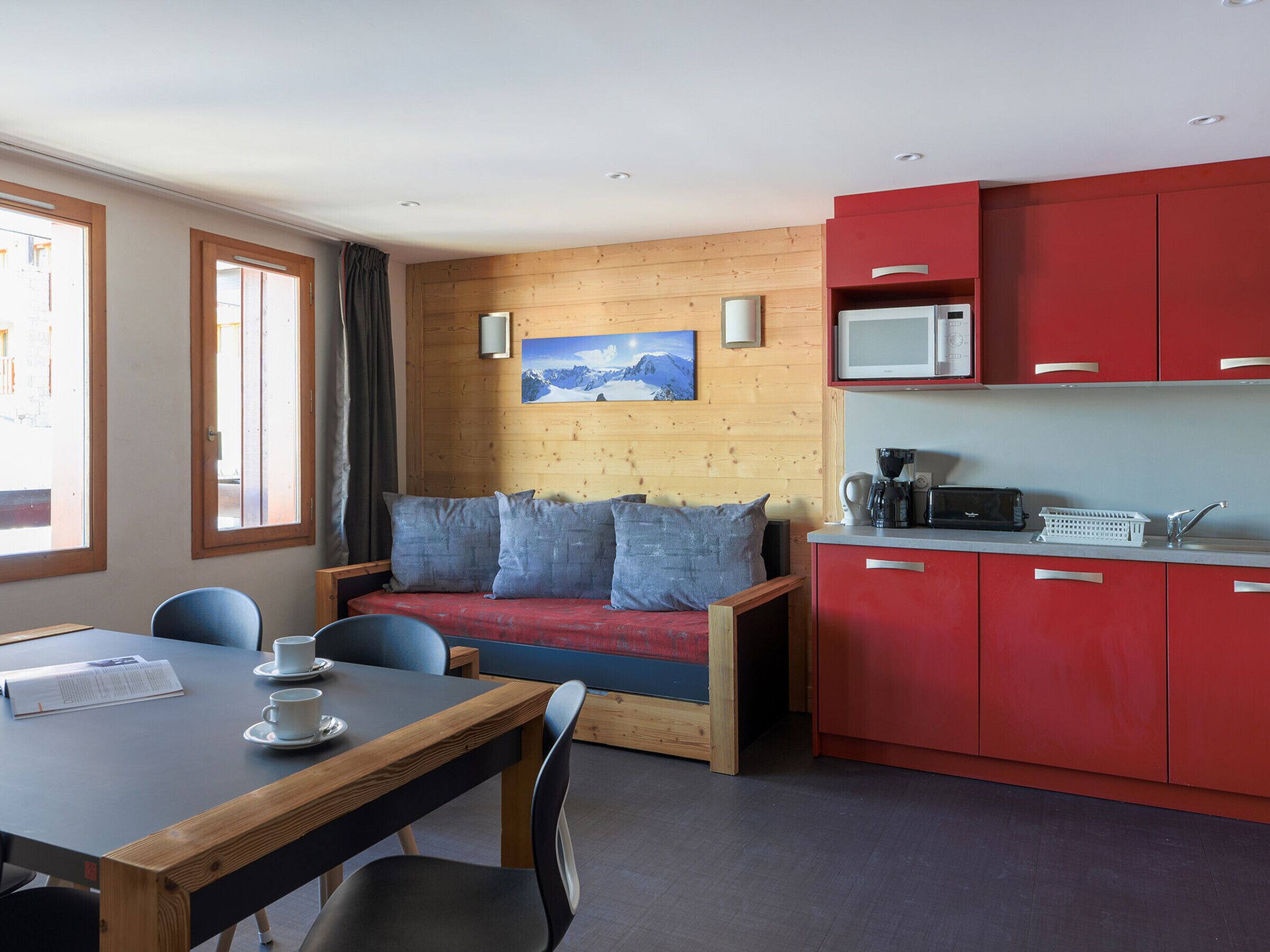 A compact room with a wooden wall, a red cabinet, a wooden table with chairs, a bed with blue pillows, and a painting of a mountain.