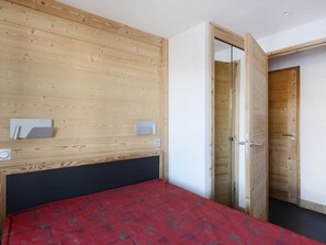 A bedroom with a wooden headboard, a red bedspread, a bedside table, and a mirror. There is a door leading to another room. - Spacious 3-room apartment, sleeps 6, pets allowed (La Plagne Montchavin Les Coches)