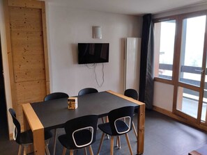 A room with a wooden table, black chairs, a television mounted on the wall, and a window with curtains. - Spacious 3-room apartment, sleeps 6, pets allowed (La Plagne Montchavin Les Coches)