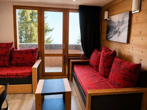 Ett rum med trägolv, en soffa med röda kuddar, ett litet bord och ett fönster med utsikt över träd.