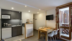 Une cuisine compacte dotée d’appareils électroménagers modernes, un coin repas avec une table et des chaises en bois, et un téléviseur fixé au mur.