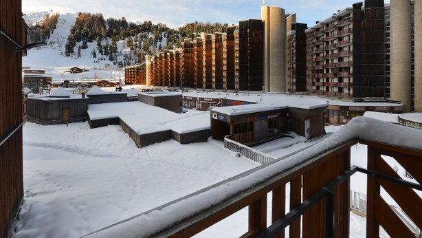 Une station de montagne enneigée comprenant plusieurs bâtiments, dont un grand hôtel doté de balcons, et un télésiège visible au loin.
