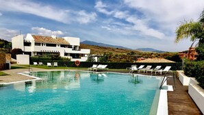 Una piscina nítida amb gandules blanques i un bou de salvament, envoltada d'una terrassa de fusta i un edifici blanc amb teulada d'aquarella.