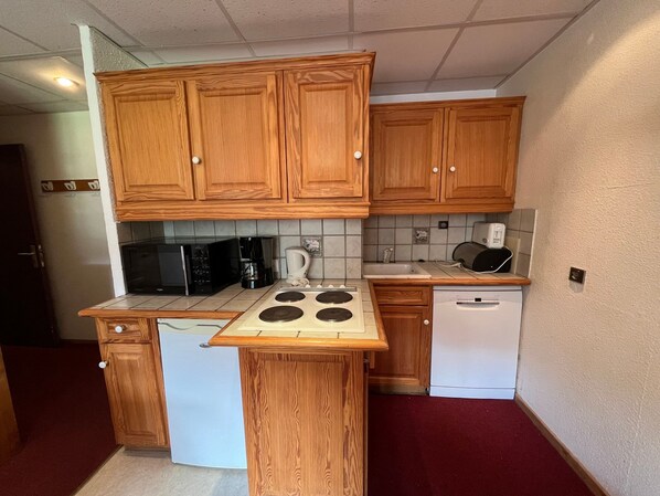 A compact kitchen with wooden cabinets, a microwave, a coffee maker, a kettle, a toaster, a stove, and a dishwasher.