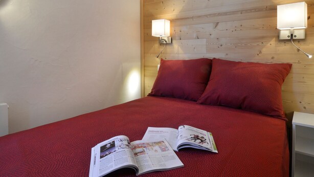 Une chambre avec une tête de lit en bois, deux lampes de lecture fixées au mur, une couverture rouge et un magazine sur le lit.