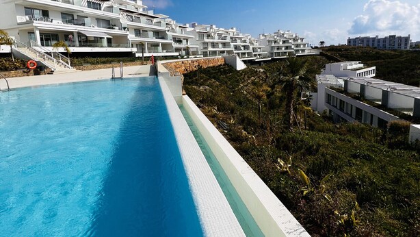 En moderne resort med en lang swimmingpool, hvide bygninger og en klar blå himmel.