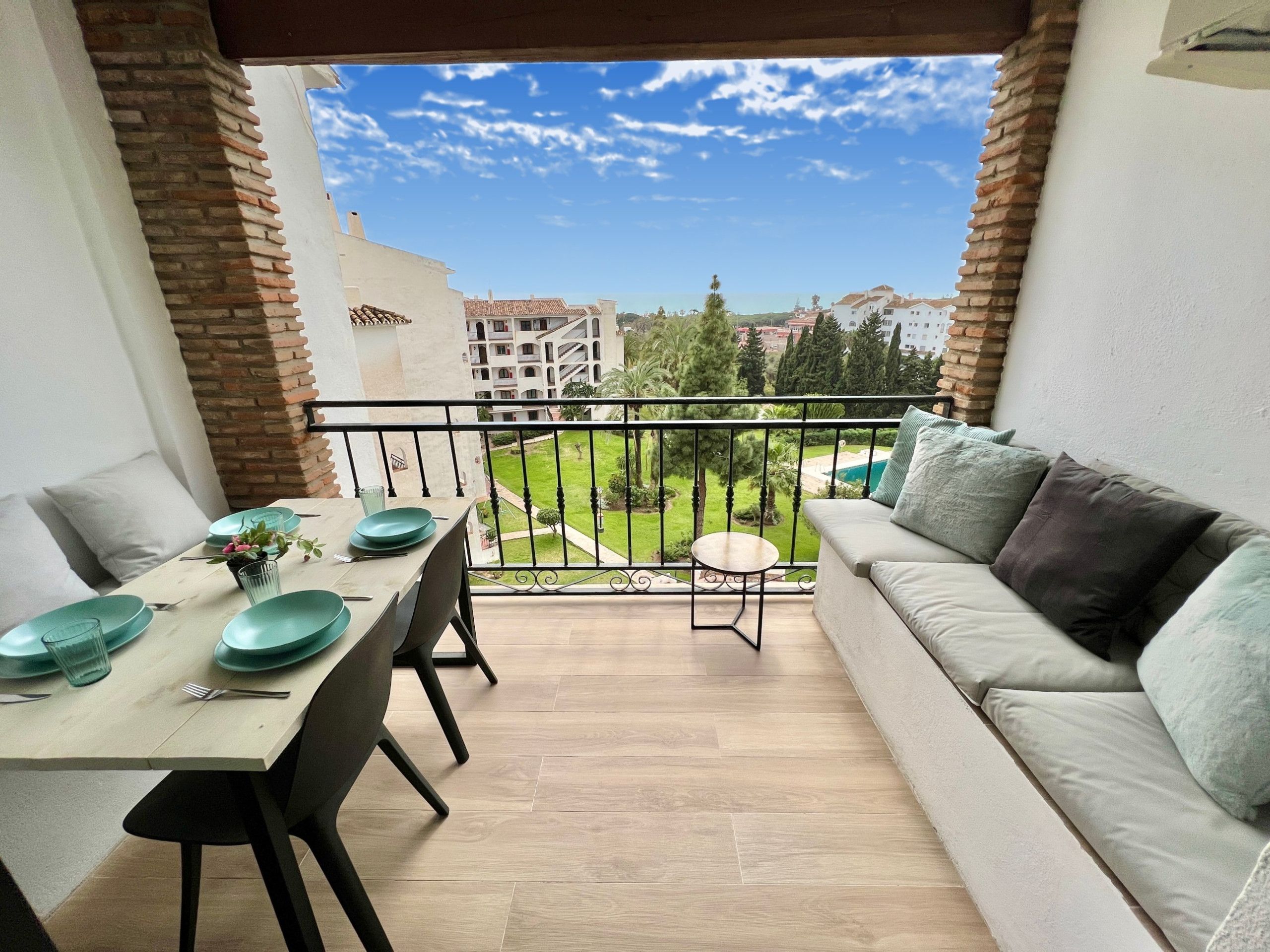 A balcony with a dining table set for two, a sofa, and a view of a residential area.