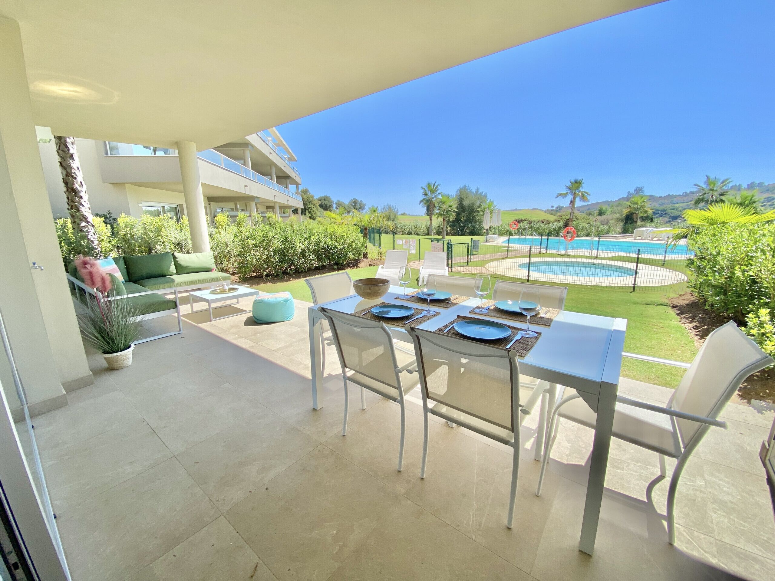 Pátio externo com uma mesa de jantar preparada para quatro pessoas, rodeado por cadeiras brancas. Um piscina ao fundo com uma cerca, e um edifício com varandas visíveis.