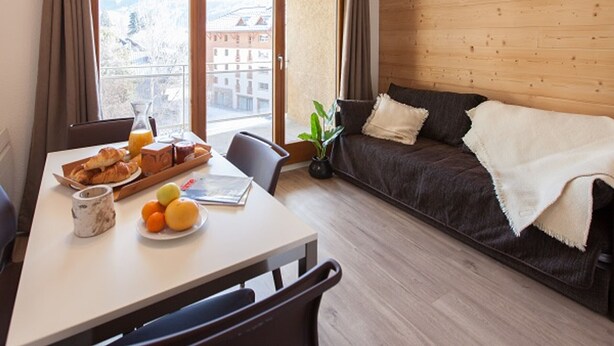 Un salon moderne avec un mur en bois, un canapé, une table à manger avec des aliments pour le petit-déjeuner, et une vue sur les bâtiments à travers de grandes fenêtres.