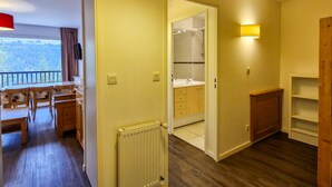 A room with a wooden floor, a dining table with chairs, a radiator, a bathroom with a mirror and sink, and a view of the outdoors. - Bright apartment with balcony in the station center (Flaine)