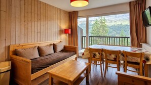 A cozy room with a wooden sofa, a dining table with chairs, a red lamp, and a large window with a view of a forest and mountains. - Bright apartment with balcony in the station center (Flaine)