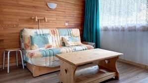Una habitación acogedora con pared de madera, un sofá con cojines florales, una mesita de madera y una ventana con cortinas.