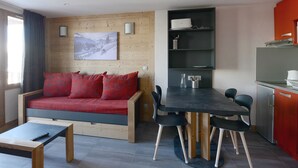 Ein modernes Wohnzimmer mit einem roten Sofa, einem Esstisch mit Stühlen, einer Küchenzeile inklusive Kühlschrank und einer Holzwand mit einem gerahmten Bild.