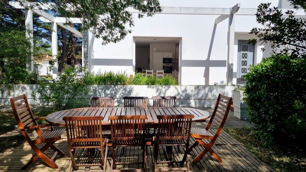 Eine Holzterrasse mit rundem Tisch und Stühlen, umgeben von Grünpflanzen und einem Gebäude mit weißen Kacheln und einem Ziegeldach.