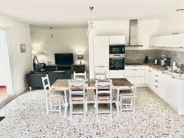 Een kleine keuken met witte kastjes, een houten eettafel en een televisie op de achtergrond.