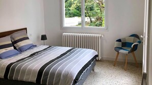 Ein Schlafzimmer mit Bett, Stuhl, Heizkörper und einem Fenster mit Blick auf Grünflächen.