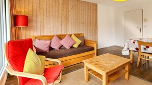 Un salon chaleureux avec un canapé en bois, un fauteuil rouge, une table basse en bois et une table à manger recouverte d’une nappe à motifs.