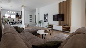 A modern living room with a large sectional sofa, a round coffee table, and a flat-screen TV mounted on a wooden panel. - Villa Dora (Policnik)
