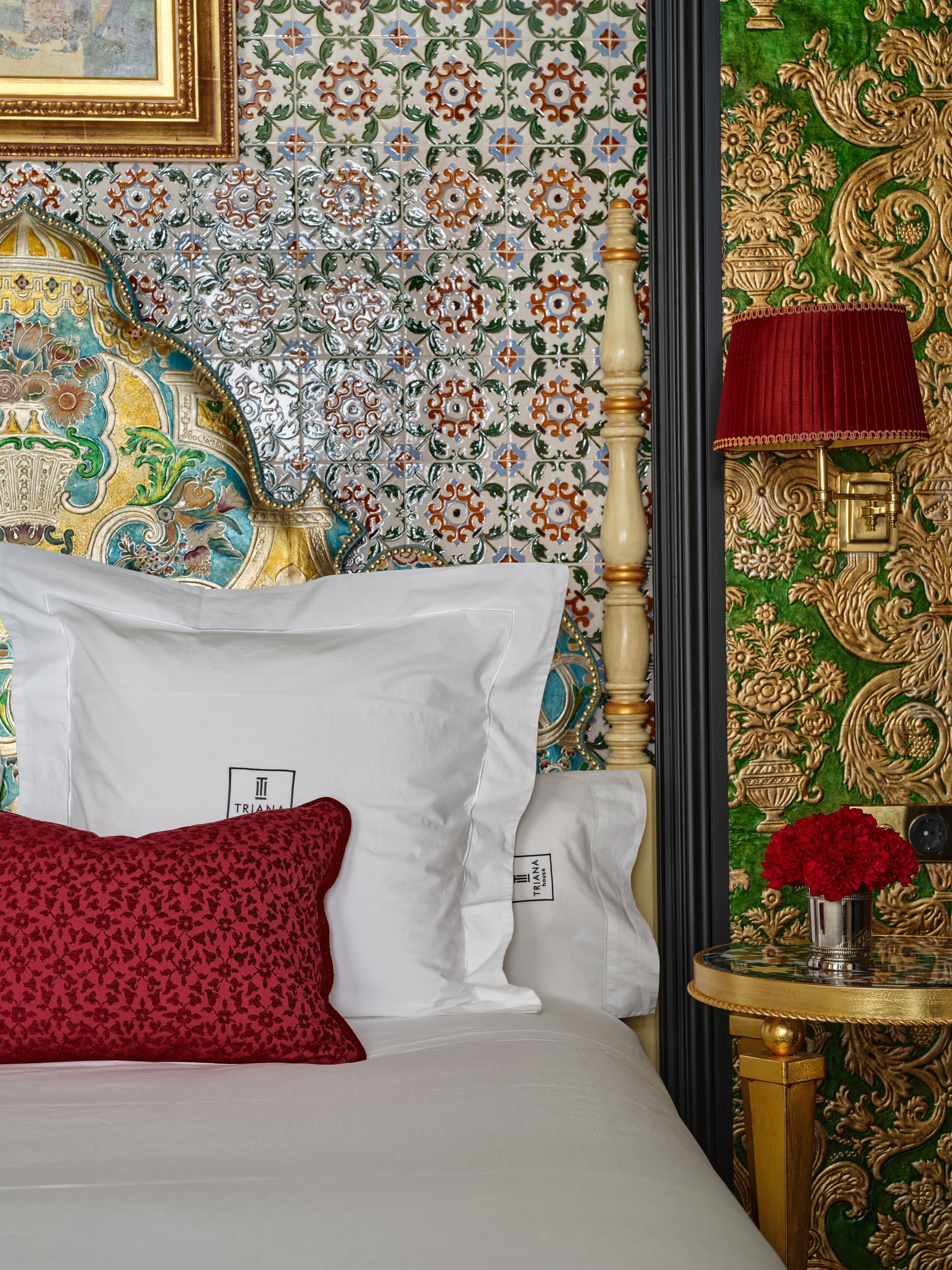 A bed with white and red pillows, a red patterned pillow, a gold-framed headboard, and a small table with a red lamp and a vase of flowers.